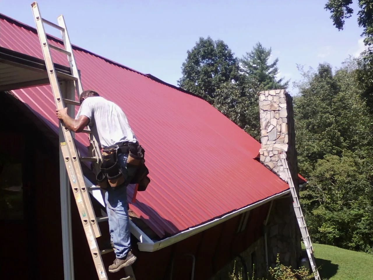 Roofer on a ladder beside a red metal roof and stone chimney — Soffit & Fascia Repair by Roofing Brothers in Kings Mountain